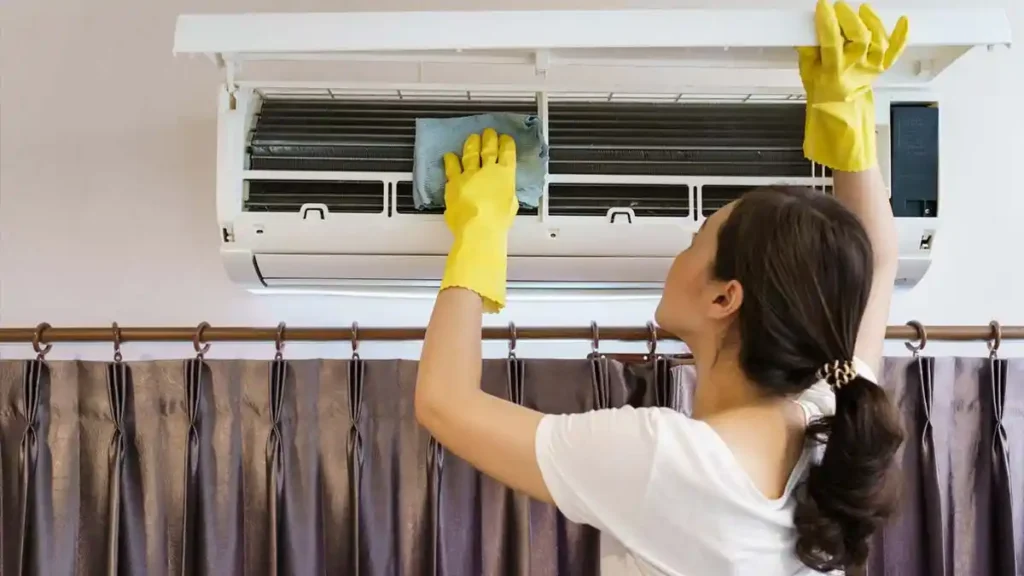 امرأة ترتدي قفازات واقية وتقوم بمسح الأجزاء الداخلية لمكيف الهواء بقطعة قماش، مما يوضح الطريقة الصحيحة لتنظيف نظام التكييف المنزلي بأمان.