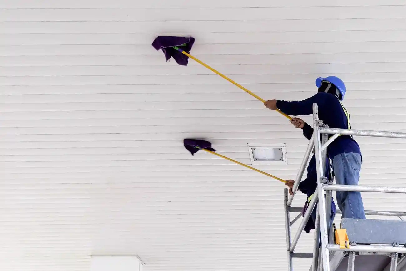 تنظيف الأسقف العالية في قصر واسع باستخدام أدوات تنظيف طويلة، مع ظهور تراكم الغبار على الأسطح العلوية بسبب المساحات الكبيرة وارتفاع السقف.